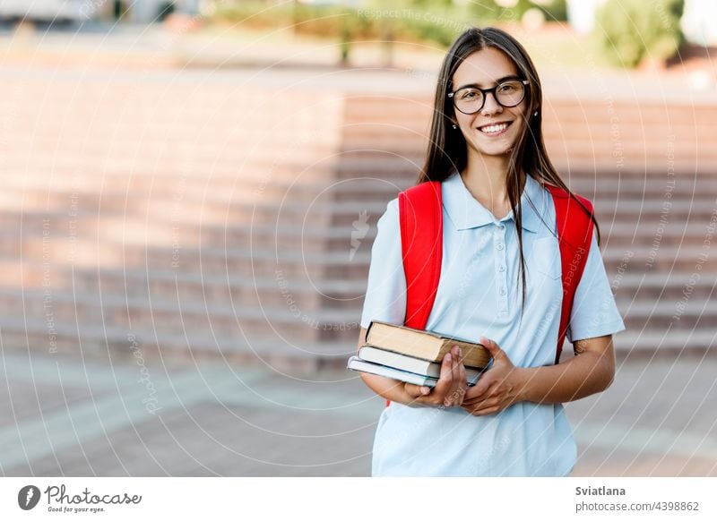 Eine lächelnde Studentin mit Brille, Büchern und einem roten Rucksack. Porträt einer Studentin vor einem städtischen Hintergrund. Das Konzept der Bildung