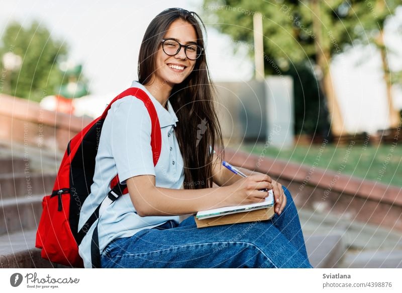 Porträt einer charmanten Studentin mit Brille, die auf einer Treppe in der Stadt sitzt Schüler Sitzen schreibend Mädchen Buch Notebook Rucksack fokussiert