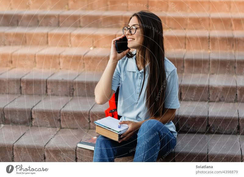 Ein fröhliches Mädchen spricht mit ihrer Freundin über ein Handy und sitzt mit einem Notizbuch auf der Treppe des Campus Lernen reden Lächeln Rucksack Buch