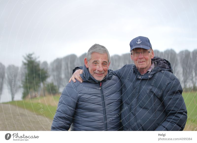 Freunde - Porträt von zwei freundlichen Senioren in Winterjacken draußen in der Natur Mensch Mann Jacke Kappe grauhaarig Bart Brille Landschaft Baum Weg Lächeln