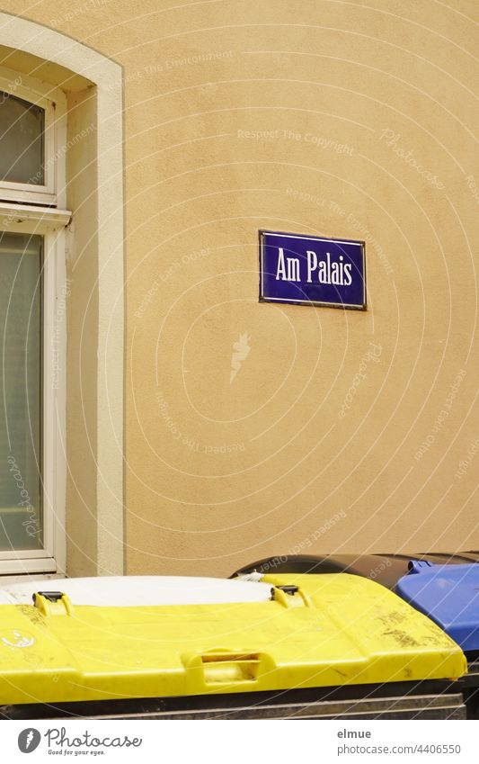 lila Straßennamenschild - Am Palais - an einer beigefarbenen Hauswand und zwei Müllcontainer vor dem Fenster / wohnen Straßenschild