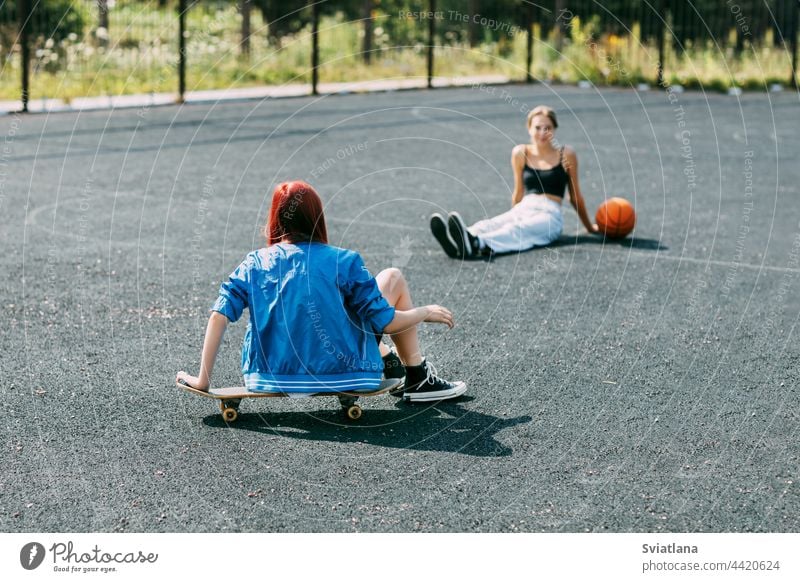 Ein junges Mädchen sitzt auf einem Skateboard im Freien auf einem Basketballplatz mit ihrem Freund, der Basketball spielt Skateboarderin Sport Skateboarding