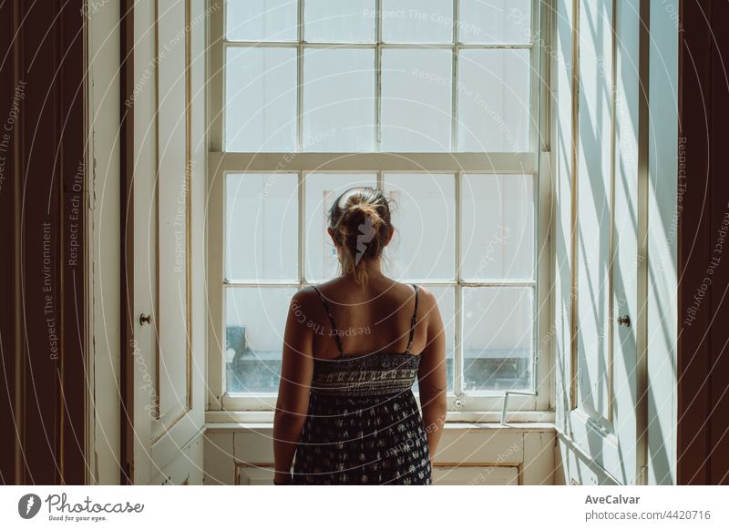 Junge Frau, die ein Kleid trägt und durch ein Fenster schaut, mit dem Rücken zur Kamera, Stress- und Angstkonzepte, dunkles Bild, Traurigkeit Einsamkeit
