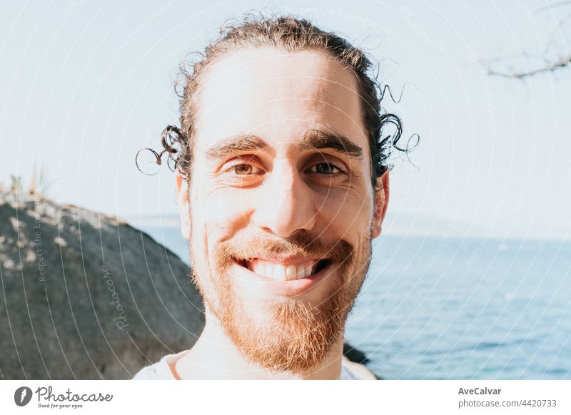 Super close up Porträt eines jungen Mannes mit einem leeren weißen Hemd lächelnd in die Kamera an der Küste von Spanien während eines Strandtages, psychische Gesundheit Glück und Zukunft Konzept