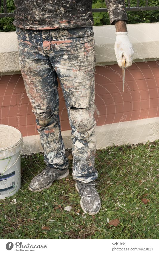 Stehender Maler vor frisch angestrichener Mauer Hose Schuhe Pulli Handschuh und Werkzeug mit reichlich Farbflecken Wandfarbe Unterkörper Beine stehen Farbeimer