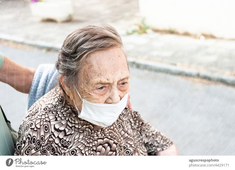 Nahaufnahme einer achtzigjährigen Frau, die in einem Rollstuhl die Straße hinuntergeht, mit Blick nach vorn in einem gemusterten Hemd, die eine Schutzmaske trägt.