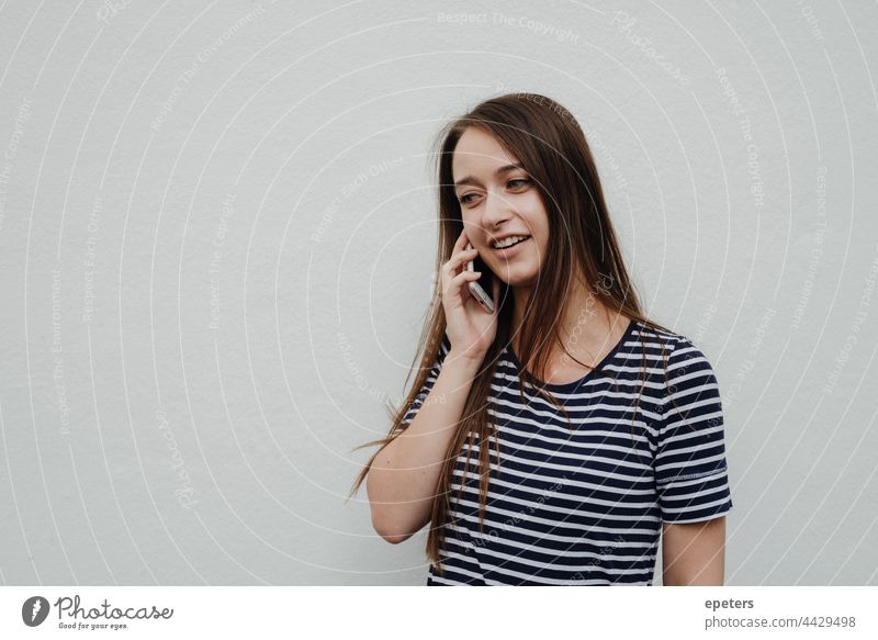 Junge Frau hält sich ein Smartphone ans Ohr und steht vor einer weißen Wand braune Haare Freizeitbekleidung Textfreiraum gen z Nachrichten erhalten