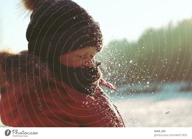 Schneeflocken glitzern im Sonnenlicht in der Luft vor dem Gesicht eines Kindes, das seine Augen geschlossen hat. Ein Junge in Winterkleidung spielt an einem frostigen, sonnigen Wintertag im Freien mit Schnee.