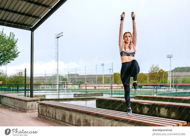 Junge Frau trainiert mit Hanteln im Freien Training Kurzhantel Arme hoch strecken Bein Knie Textfreiraum Kraft Übung Aufwärmen jung auf einer Bank Gesundheit