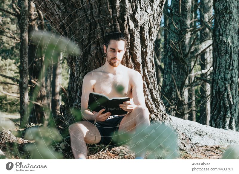 Ein Mann in Stämmen shirtless Lesen eines Buches gegen einen Baum während eines Sommertages, entspannen und chillen Konzepte, gutes Leben, junge Menschen
