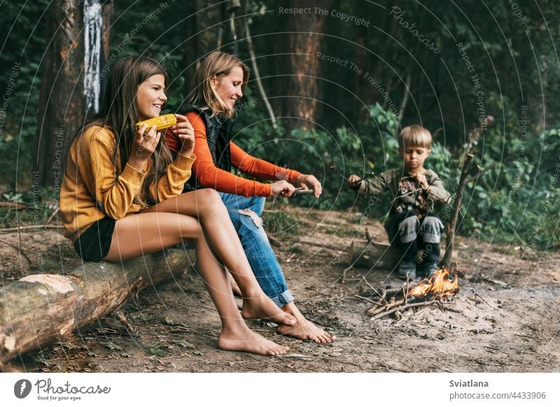 Eine junge Mutter sitzt mit ihren Kindern bei einem gemeinsamen Urlaub am Lagerfeuer, ein Mädchen isst Mais Camping Mama Wochenende Wanderung Tochter Sohn