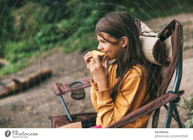 Ein junges Mädchen in einem leuchtend gelben Pullover sitzt in einem Campingstuhl am Ufer des Sees und isst Mais Lager Stuhl Sitzen essen Frau Natur Menschen