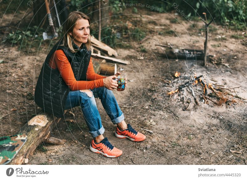 Eine junge Frau wärmt sich am Feuer mit einer Tasse wärmenden Tees im Wald trinken Sitzen Natur Tourist Abenteuer Urlaub Lagerfeuer Freudenfeuer reisen