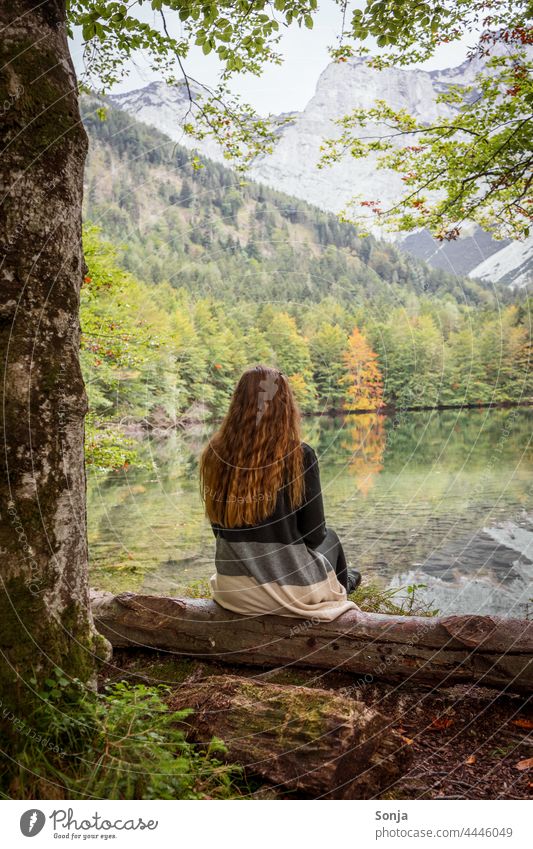 Junge Frau mit langen roten Haaren sitzt an einem See und genießt die Aussicht jung Seeufer sitzen Baumstamm Wald Herbst Rückansicht lange Haare gelockt schön