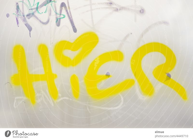 HIER steht in knallig gelben Buchstaben und mit einem Herz als i-Punkt an der grauen Metallwand / Graffito hier Ortsangabe Treffpunkt Graffiti Herzchen Schrift