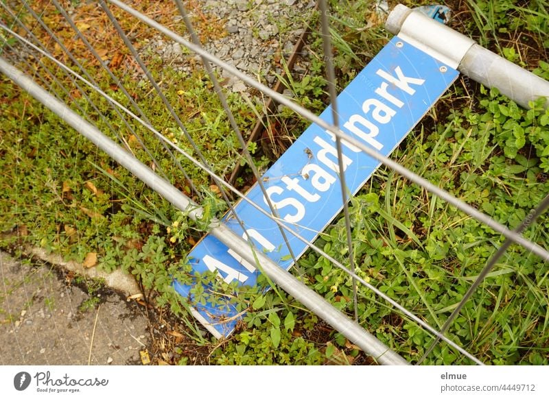 blaues Straßennamenschild  - Am Stadtpark - liegt unter einem Absperrgitter auf der Wiese Schild abgebrochen Orientierung Vandalismus Straßenschild Umbau