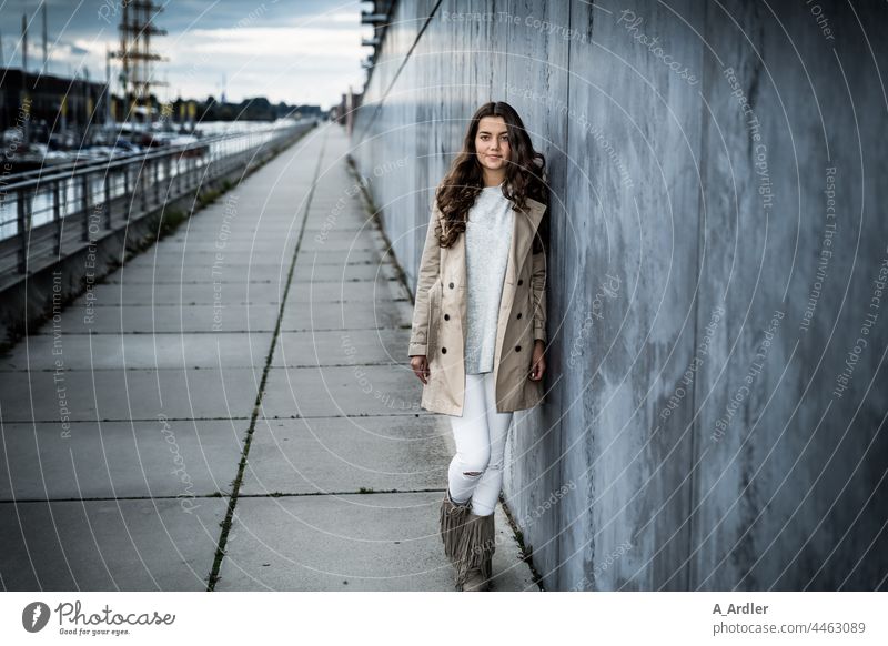 junge Frau im Mantel lehnt an einer Mauer, Hafen im Hintergrund Mädchen Junge Frau Abend abendämmerung Mensch Wand Mode Bekleidung brünett langhaarig