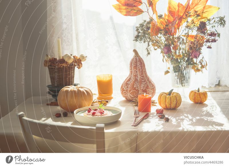 Herbstlicher Frühstückstisch mit Kürbissen, orangefarbenen Herbstblättern und Blumen, Waffeln mit Schlagsahne und Kerzen. Häusliches Stillleben am Fenster mit Sonnenlicht