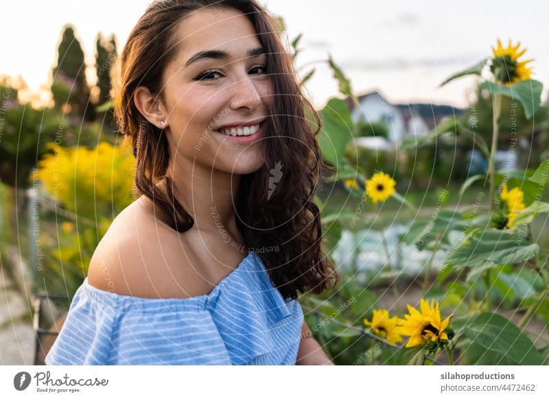 Schöne junge Frau, die durch eine Kleingartenanlage geht und dabei ihr türkisfarbenes Fahrrad mit Sonnenblumen im Korb schiebt. Frauen kaukasische Ethnizität