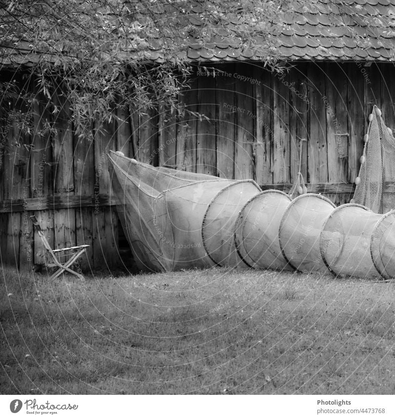 Ein Fischernetz hängt an einer Holzhauswand, daneben liegt eine Aalreuse auf der Wiese mit einem Klappstuhl daneben. Reuse Schwarzweißfoto Netz