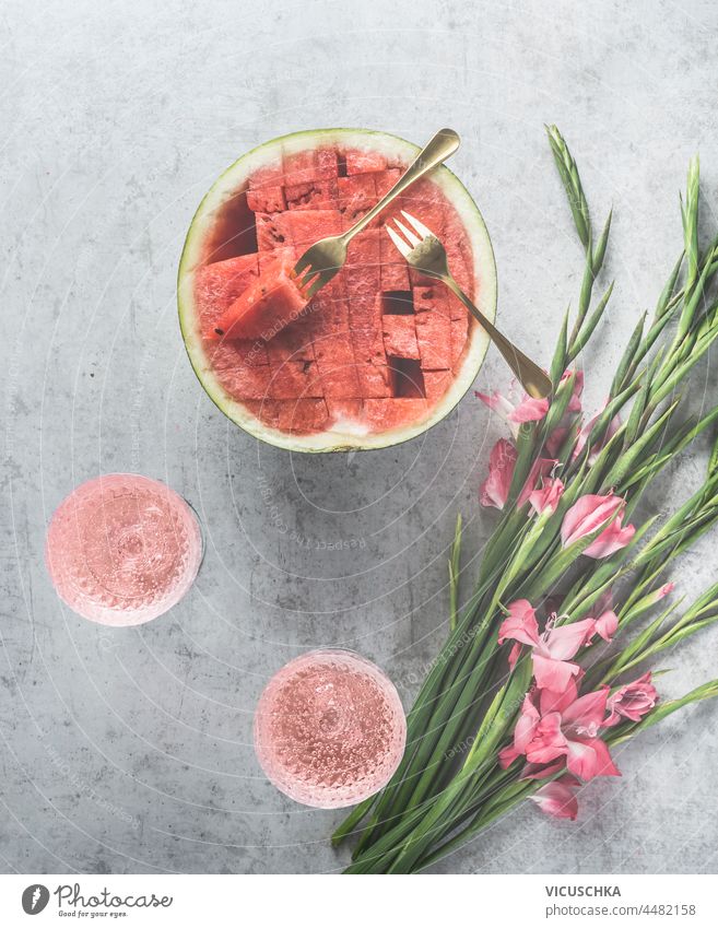 Wassermelonen-Cocktails mit Sekt in Weingläsern, geschnittener Wassermelone und rosa Blumen auf hellgrauem Beton-Küchentisch. Sommerfest mit hausgemachten Getränken und Obst. Ansicht von oben.