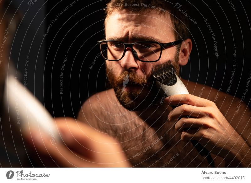 Ein junger Mann rasiert und stutzt seinen Bart mit einem elektrischen Rasierapparat, im Badezimmer zu Hause, früh am Morgen, um sich vor dem Ausgehen zu rasieren