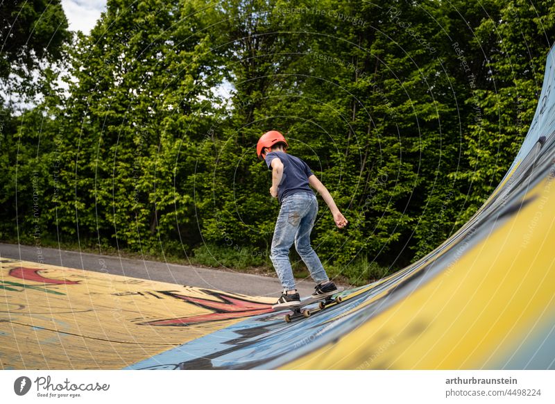 Junge in Jeans und mit Helm fährt auf Skateboard in der Halfpipe im Skatepark im Freien Tageslicht asphalt bewegung boardsport cool deck draussen freizeit