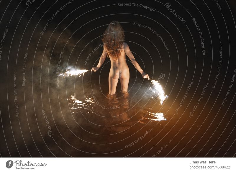 Ein wildes und nacktes Mädchen schwimmt in der Nacht. Erotisches Bild von einem brünetten Modell. Feuerwerk in ihren Händen, sexy Rücken zeigen ihre perfekten Kurven, Rauch und dunkles Wasser macht diese Szene stimmungsvoll und sinnlich.