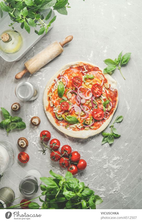 Hausgemachte Pizza mit Zwiebeln, Salami, Tomaten und Basilikum auf grauem Küchentisch mit hölzernen Nudelholz, Pilzen und Küchenutensilien. Zubereitung von italienischem Essen zu Hause mit frischen Zutaten. Ansicht von oben.