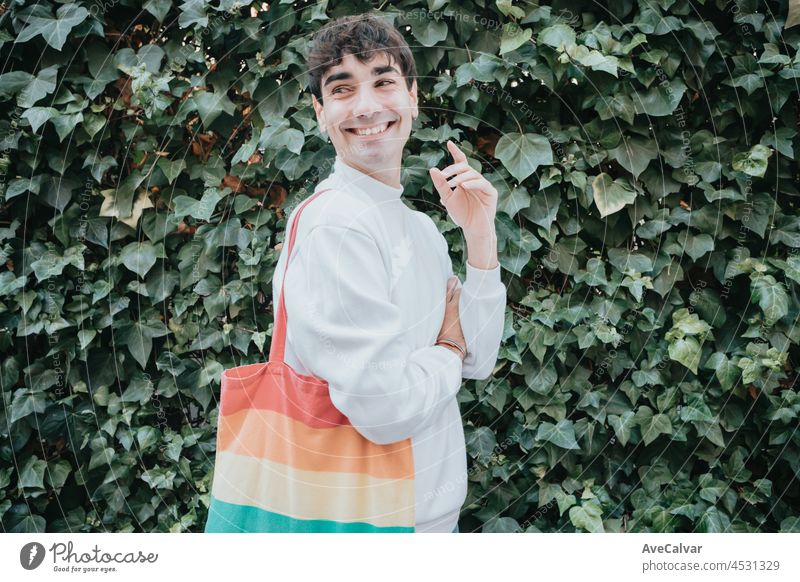 Porträt eines jungen lgbt Mann mit einer Tragetasche von Regenbogen Stolz Flagge lächelnd weg von der Kamera, Stadt Homosexuell Stolz Lebensstil, beste Zeit des Tages, soziales Netzwerk