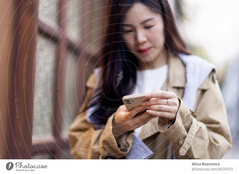 Stilvolle junge asiatische Frau in Freizeitkleidung, die beim Posten von Fotos in sozialen Medien auf den Handybildschirm schaut und über interessante Texte nachdenkt. Lifestyle-Bloggerin überprüft Blog-Kommentare im Freien im Herbst