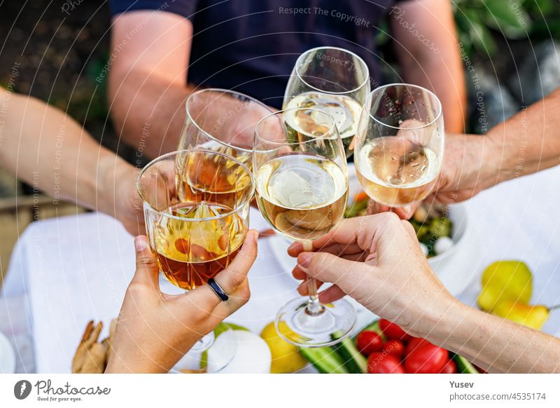 Eine glückliche Familie, die bei einem sommerlichen Gartenfest zu Abend isst und auf die Getränke anstößt. Es wird angestoßen und getrunken. Festliches Familienessen im Hinterhof. Männliche und weibliche Hände halten Gläser mit Wein. Lebensstil