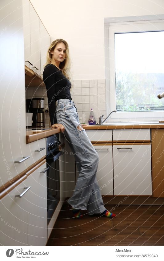 Junge, schlanke, große Frau mit langen, blonden Haaren steht in einer Küche an den Schrank angelehnt und schaut in die Kamera junge Frau langhaarig schön