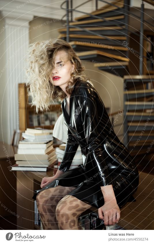 Ein wunderschönes Modemodell posiert mit ihrem schwarzen Latex-Outfit. Ihr lockiges blondes Haar sieht toll aus. Die roten Lippen sind so sinnlich. Der Hintergrund scheint eine Bibliothek zu sein.
