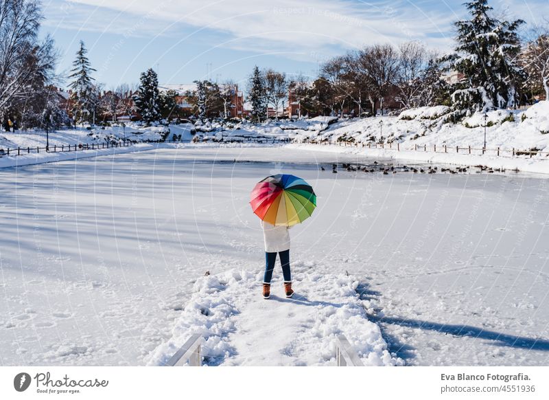 Rückansicht einer Frau, die einen bunten Regenschirm hält, der auf einem verschneiten Steg an einem zugefrorenen See steht, an einem sonnigen Tag. Winter Schnee