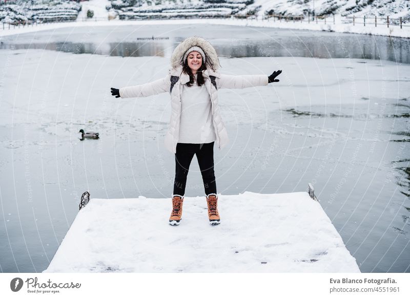 glückliche kaukasische Frau mit geschlossenen Augen und ausgestreckten Armen, die im Winter auf einem verschneiten Steg am zugefrorenen See steht. Winter Lebensstil in der Natur