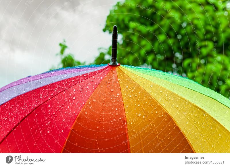 Regen auf Regenschirm in Regenbogenfarben unter starkem Regen gegen Hintergrund des bewölkten Himmels. Regenwetter-Konzept. herbst hintergrund schlecht blau