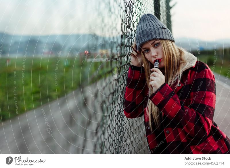 Junge Frau mit Wollmütze lehnt an einem Zaun und raucht Porträt jung Rauchen vaping alternativ in die Kamera schauen attraktiv Verdampfer schön blond Schlag