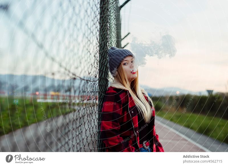Junge Frau mit Wollmütze lehnt an einem Zaun und raucht jung Rauchen vaping alternativ in die Kamera schauen attraktiv schön blond Schlag Kaukasier Zigarette