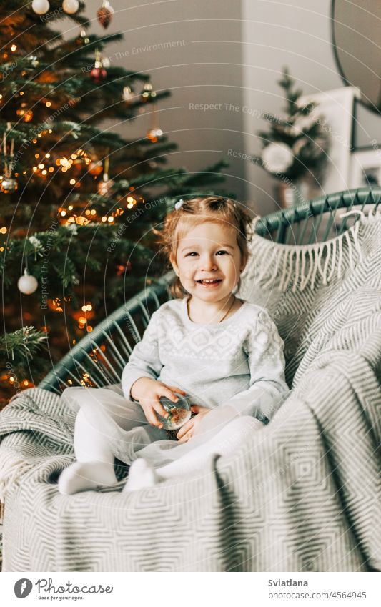 Ein kleines Mädchen sitzt in einem Sessel und lächelt vor dem Hintergrund eines Weihnachtsbaums in der Silvesternacht. Weihnachtsstimmung, Erwartung von Magie
