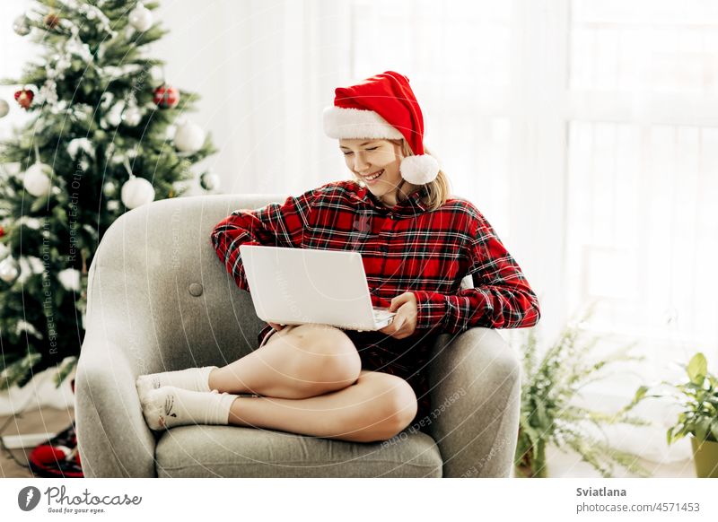 Glückliches Teenager-Mädchen mit Weihnachtsmannmütze und Laptop in einem weihnachtlich dekorierten Zimmer, das sich mit Freunden und Verwandten unterhält
