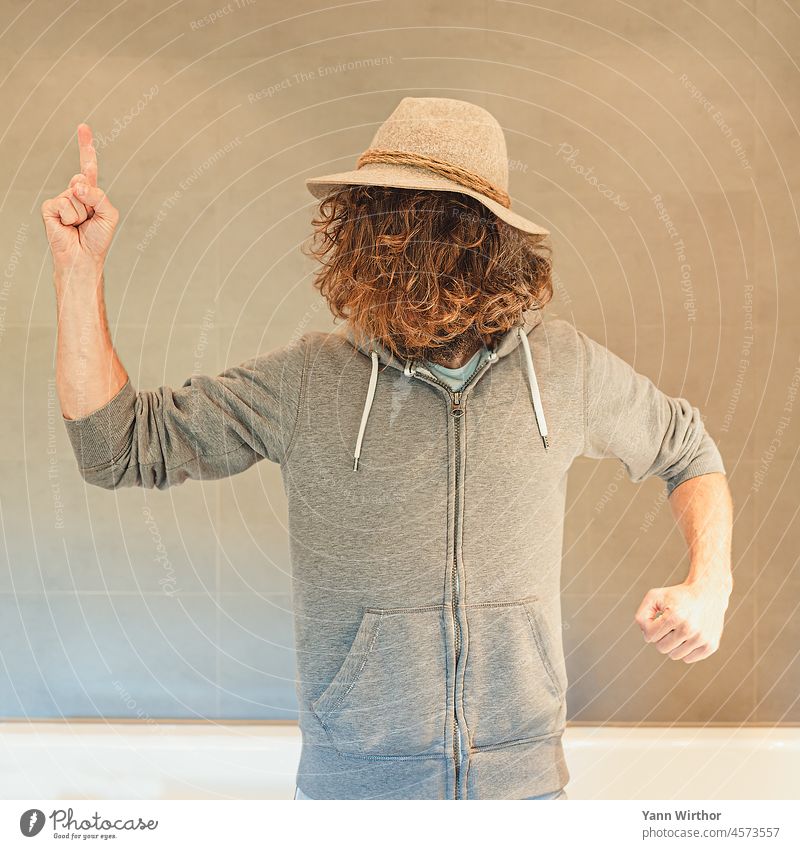 Mann mit Hut und langen lockigen braunen Haaren im Gesicht zeigt mit dem Finger nach oben Haare im Gesicht lange Haare Gesichtspflege Jacke Entschlossenheit