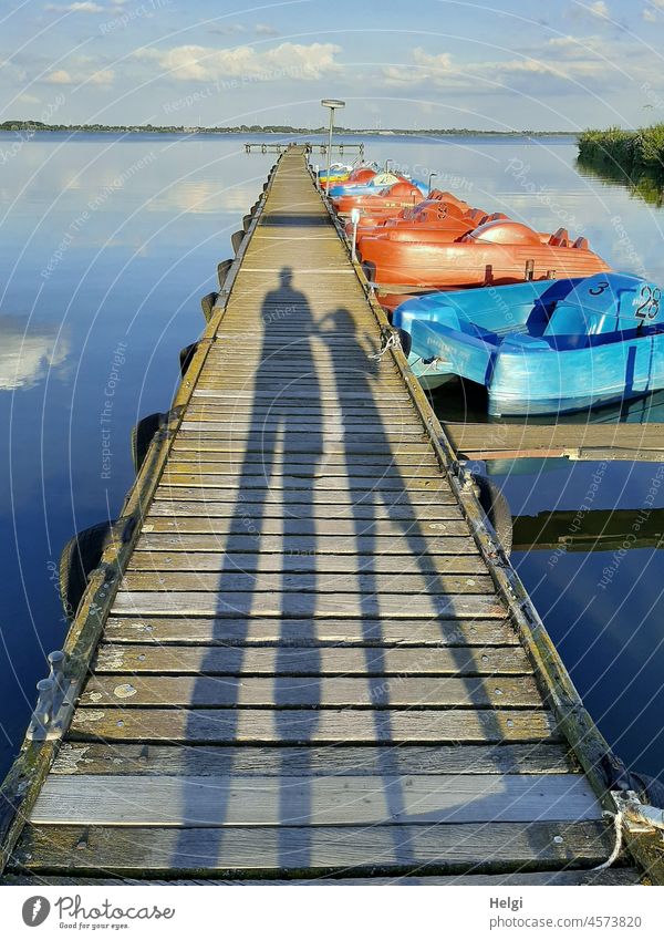 lange Schatten von zwei Menschen auf einem Bootssteg mit Tretbooten am See Frau Kind Steg Holzsteg Dümmer See Wasser Himmel Spiegelung Sonnenschein Sonnenlicht