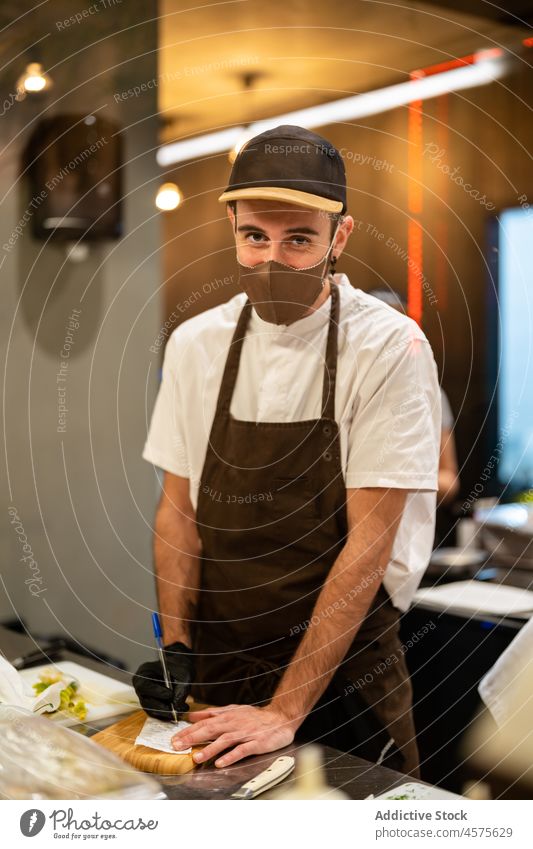 Koch schreibt auf Quittung Rechnung in Küche Mann prüfen Orden schreiben Restaurant COVID Mundschutz männlich Uniform COVID19 modern Arbeit Personal Bund 19