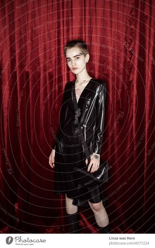 Ein wunderschönes Modemodell posiert mit ihrem schwarzen Latexkleid. Schaut direkt in eine Kamera. Mit perfekt sitzenden Tattoos. Rote Vorhänge sorgen für ein wenig Geheimnis in diesem Bild.