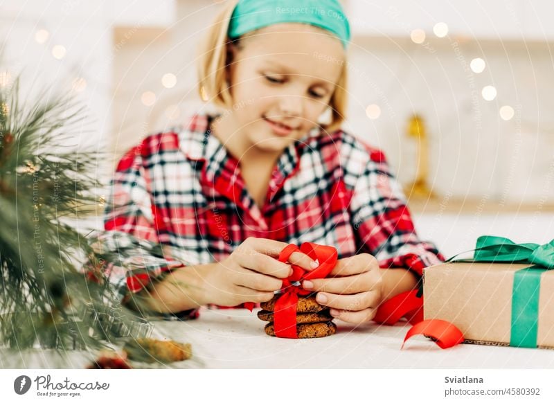 Ein hübsches Mädchen dekoriert Plätzchen mit einer leuchtend roten Schleife und bereitet eine süße Leckerei für Gäste an Weihnachten oder Silvester vor Geschenk