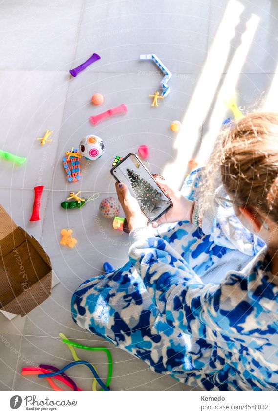 Kind, das mit Spielzeug sitzt und ein Smartphone mit einem Foto des Weihnachtsbaums benutzt Feiertag Anzeige heimwärts Weihnachten Funktelefon Mädchen feiern