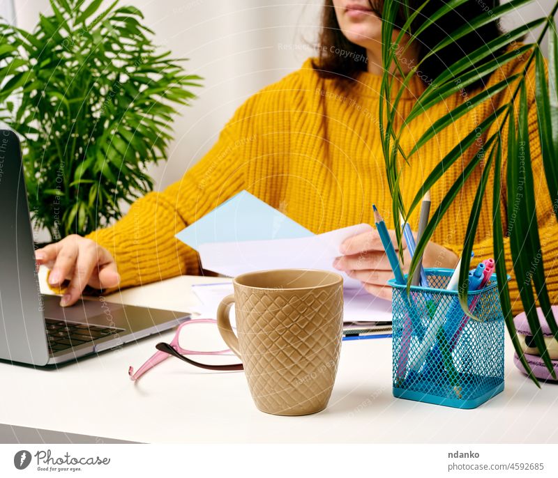 eine Frau in einem orangefarbenen Pullover sitzt an einem weißen Tisch und arbeitet an einem Laptop. Freiberufler bei Fernarbeit, Verkäuferin im Home Office online