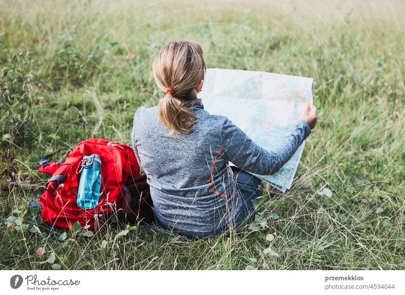 Frau mit Rucksack, die während eines Ausflugs in den Bergen eine Pause macht und auf eine Karte schaut, während sie im Gras sitzt Sommer aktiv Landkarte