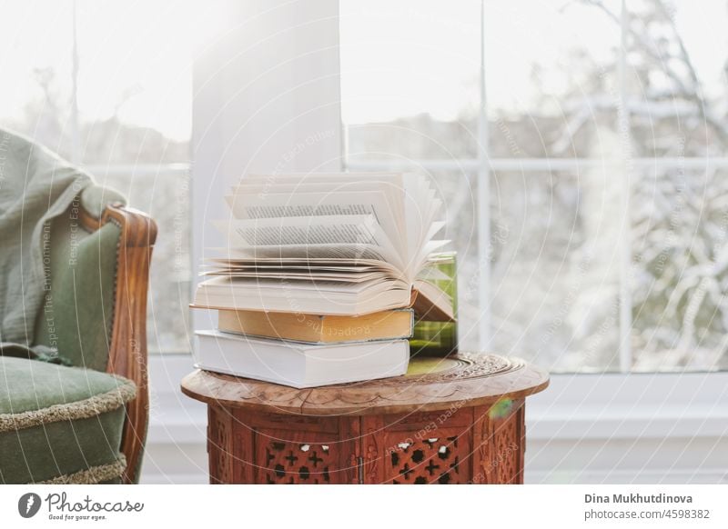 Ein Stapel Bücher auf dem hölzernen Couchtisch in der Nähe von Sessel im Zimmer mit verschneiten Bäumen Landschaft Fenster Blick. Gemütliches Zuhause im Winter.